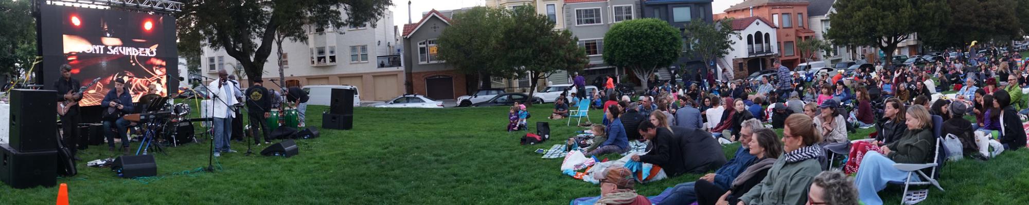 An audience of people sit on blankets at a park watching a live music performance.