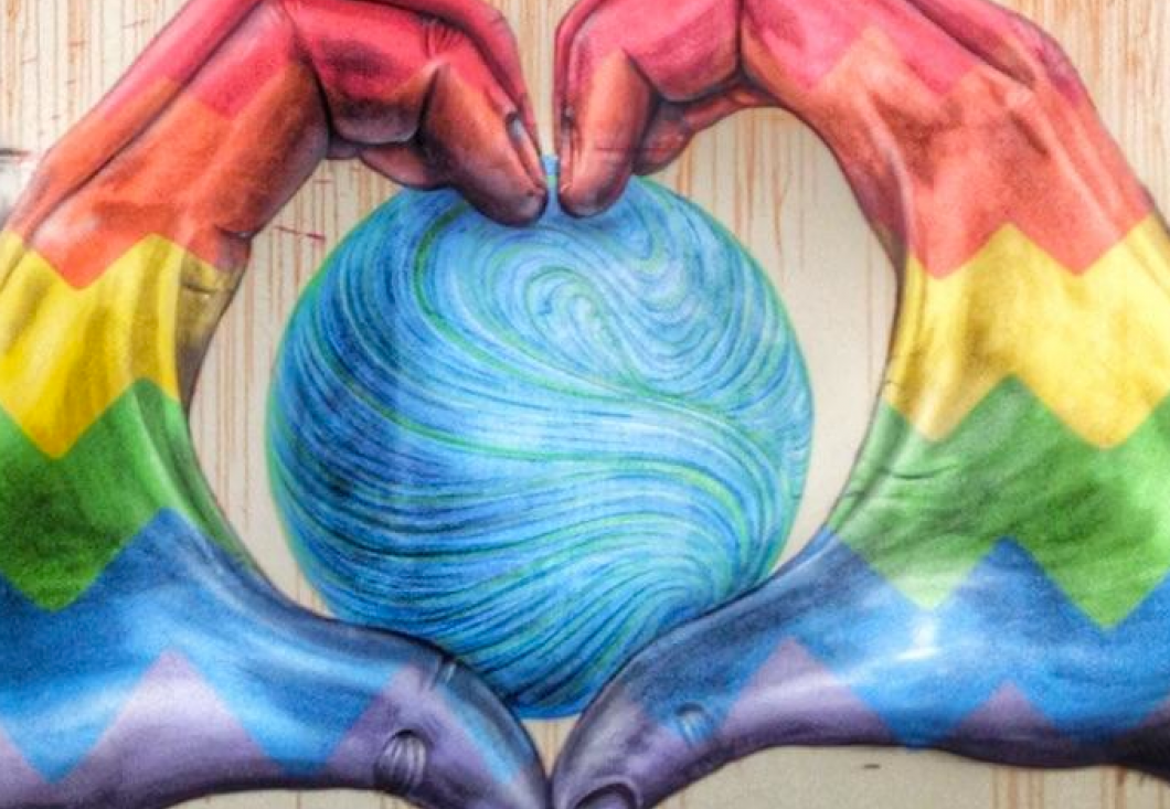 Mural on a wall in rainbow colors depicting a large sphere in blue and green. The sphere is held by two hands, grasped to display the outline of a heart with the fingers.