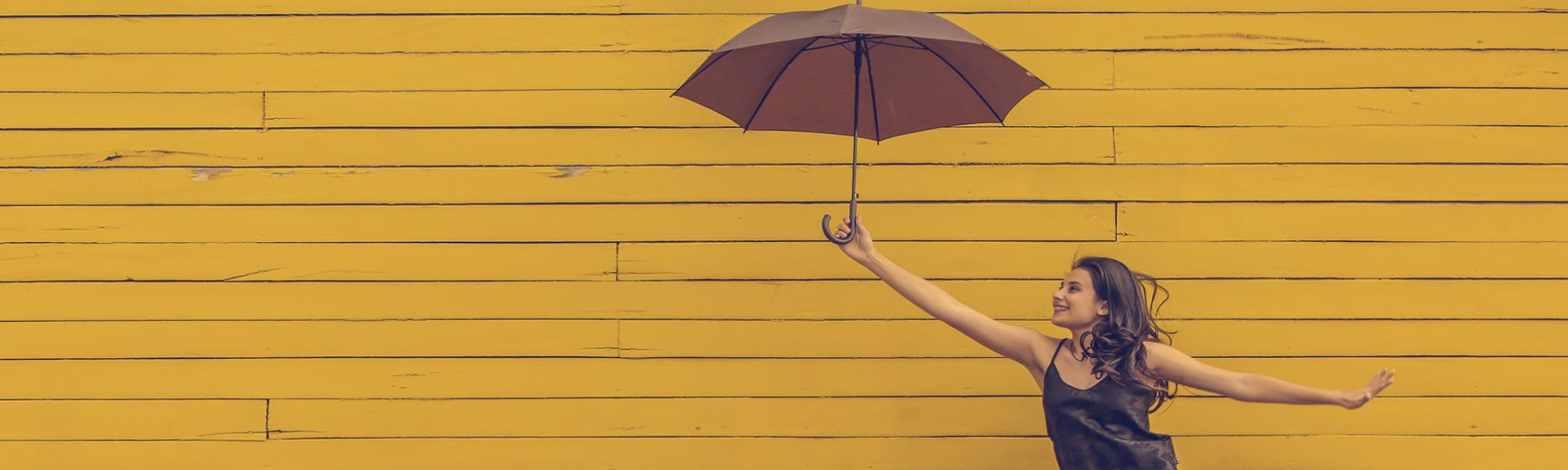 Photo of young woman in front of a bright yellow wall. She is holding an umbrella and looks like she may be blown away in flight!
